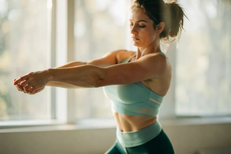 Mulher em pé, com roupas de ginástica, fazendo alongamento de braços em frente a uma janela com luz do sol