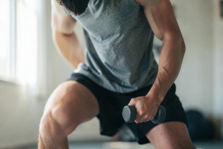 Homem com roupas esportivas demonstrando afundo com halteres em sala de estar.
