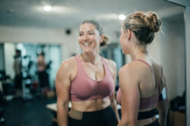 Mulher sorrindo em frente a espelho, com roupas de ginástica, em casa.