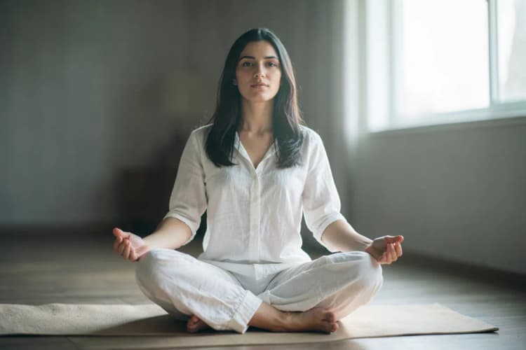 Mulher sentada em posição de lótus em um tapete de yoga, com as mãos em mudra e expressão serena.